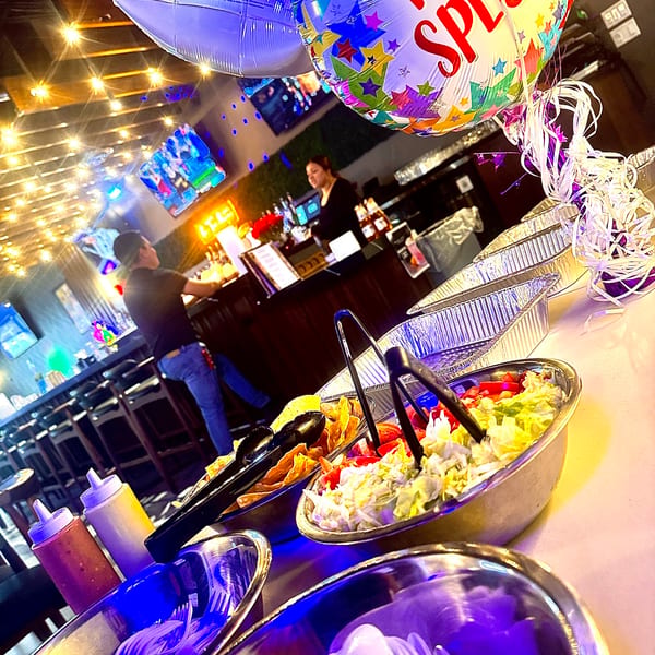 A buffet line with bowls of salad and chips, balloons, and restaurant staff in the background.