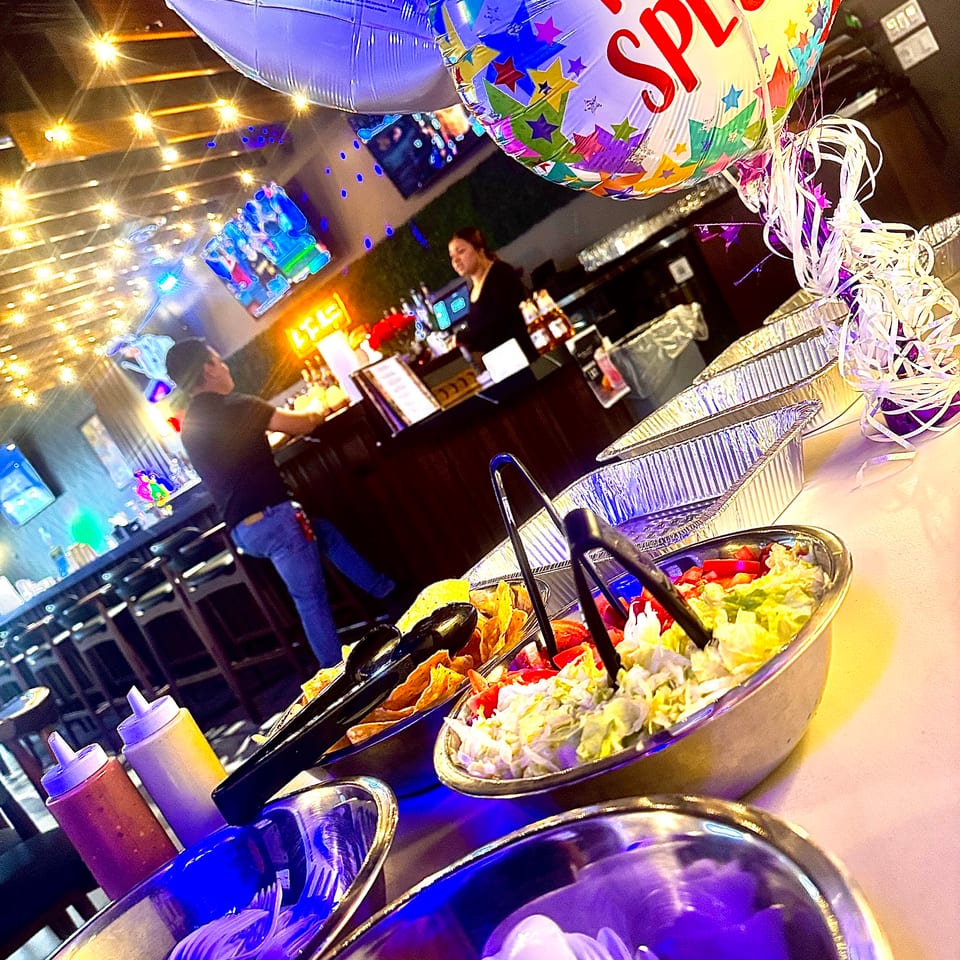 A buffet line with bowls of salad, chips, and sauces, with balloons and people in the background.