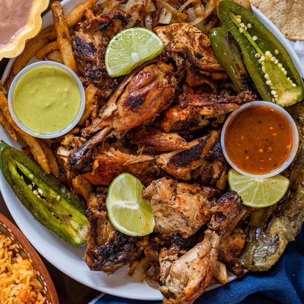 Grilled chicken pieces with fries, grilled onions, jalapeños, lime, and two sauces on a white plate.