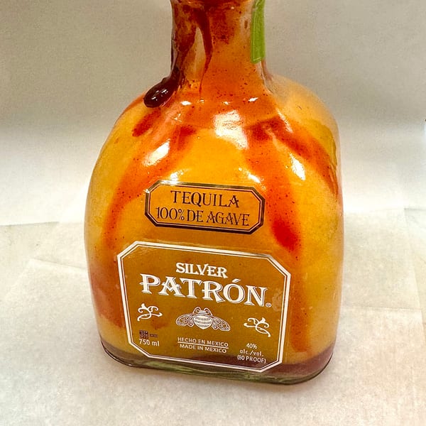 A Patron Silver tequila bottle with a dark, candy-coated straw on top, covered in red sauce.