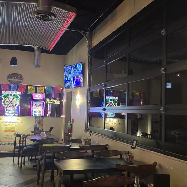 Interior of a Mexican restaurant with tables, chairs, neon signs, and a large window.