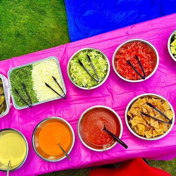 Overhead view of a vibrant pink table laden with various Mexican food toppings and sauces outdoors.