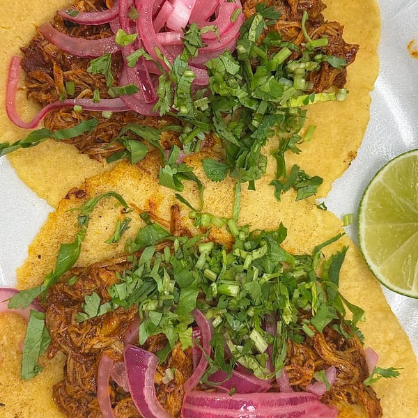 Two tacos with shredded meat, pickled red onions, cilantro, and a lime wedge on a white plate.