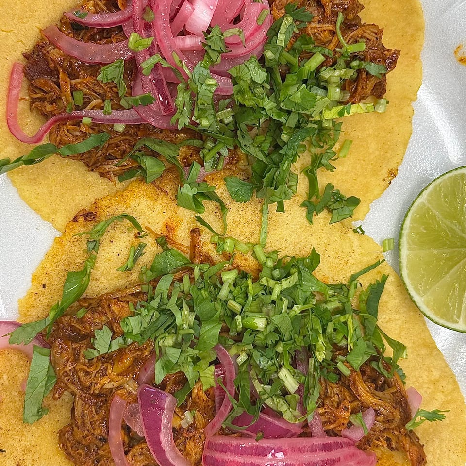 Two tacos with shredded meat, pickled red onions, cilantro, and a lime wedge on a white plate.