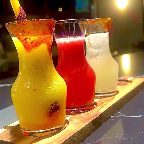Three colorful drinks, yellow, red, and white, in decorative glasses on a wooden tray.
