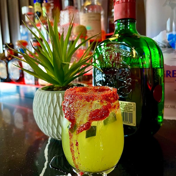 A vibrant yellow cocktail with a red chili-lime rim sits on a bar next to a potted plant and liquor bottles.