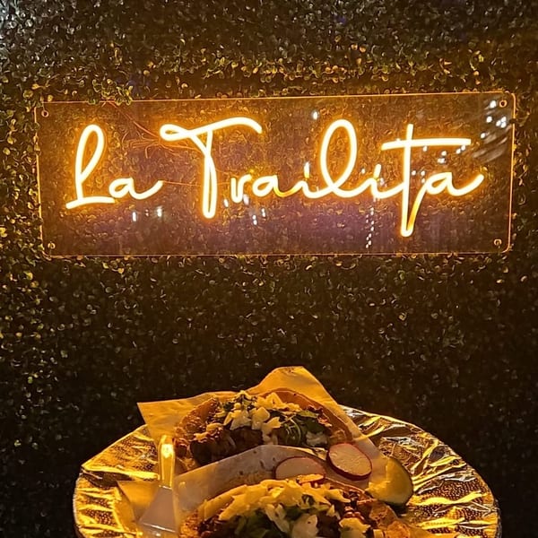 Two tacos on a foil-lined plate held up against a green wall with a neon "La Trailita" sign.