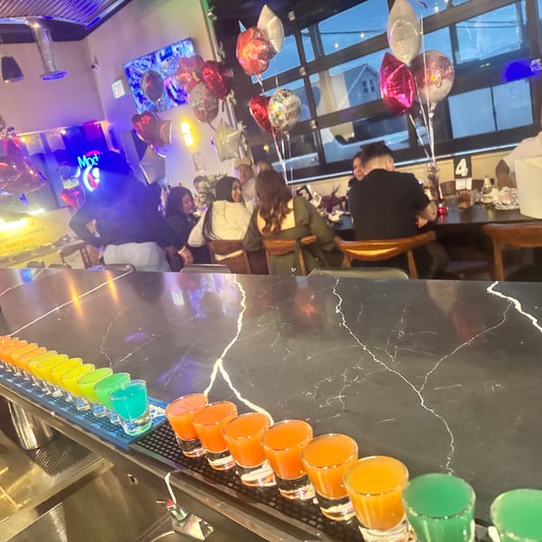 A bar counter with colorful shots in the foreground and people dining in a festive restaurant.