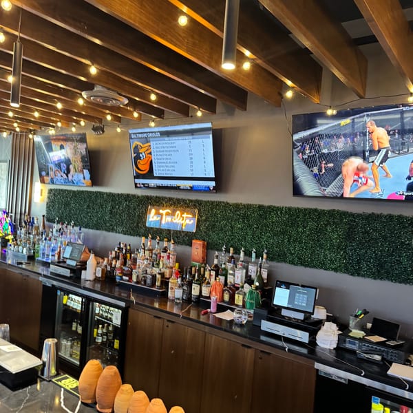 A well-stocked bar with bottles of liquor, a neon sign, and TVs showing sports, under string lights.